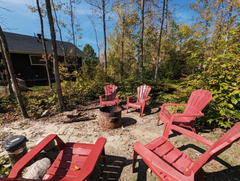 A circular arrangement of bright red Adirondack chairs surrounds a fire pit, set against a backdrop of lush greenery and a shimmering lake view.