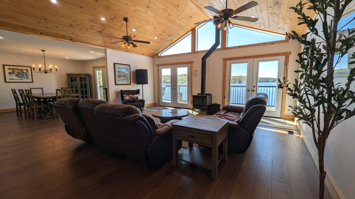 Spacious living room with vaulted wood-paneled ceilings, large windows revealing a lake view. Cozy leather seating surrounds a woodstove. Dining area in the background, perfect for gatherings.