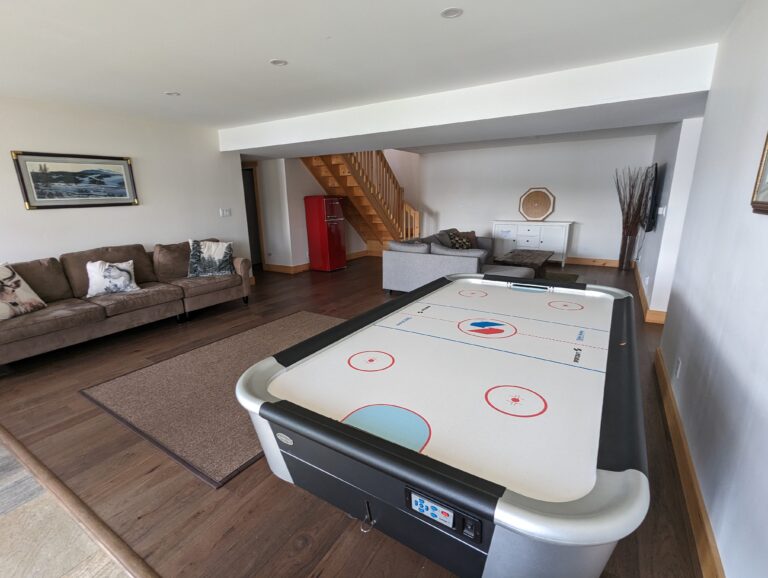 Entertainment room with an air hockey table at the center, a comfortable brown couch with throw pillows, a retro red fridge, and a wall-mounted TV. A cozy space for fun and relaxation.
