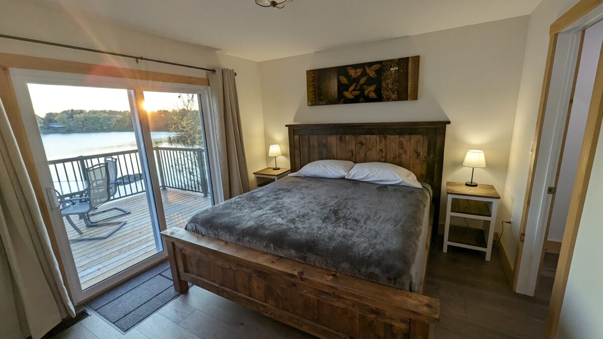 A cozy master bedroom featuring a rustic wooden king-size bed, bedside lamps, and soft lighting. A large glass sliding door opens to a private deck, offering a stunning lakeside view.