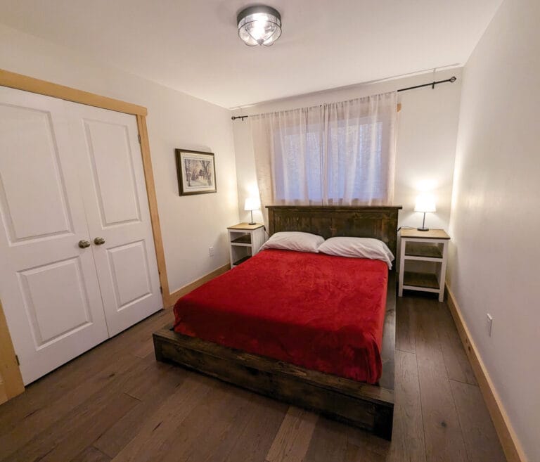 A cozy queen bedroom with a comfortable bed adorned with a red blanket. There’s a closet and side tables with lamps for added convenience and storage.