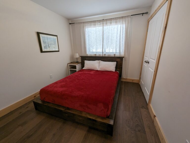 A cozy bedroom with a queen-sized bed dressed in a deep red blanket. A soft glow filters through sheer curtains. A nightstand with a lamp sits by the bed, and a closet offers storage.