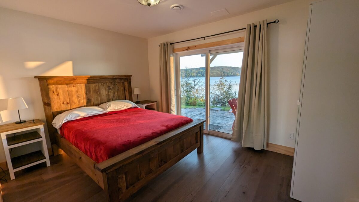 A cozy bedroom with a queen-sized bed featuring a rustic wooden headboard. The room opens up to a private deck with a picturesque view of the lake, inviting natural light to fill the space.