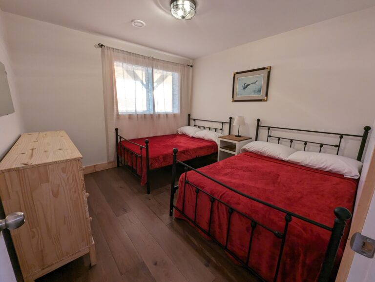 A bright room with two double beds featuring iron frames and soft, neutral bedding. A small dresser and a framed picture complete this welcoming space.